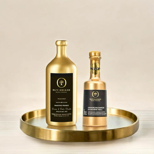 Zwei goldfarbene Flaschen, eine mit Olivenöl extra vergine und eine mit Balsamico Essig, stehen auf einem goldenen Tablett. Beide Flaschen tragen das Oliviers & Co-Logo, die linke Flasche enthält Olivenöl, die rechte Balsamico Essig.