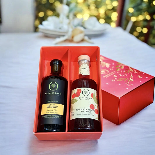 Rote Geschenkbox mit zwei edlen Flaschen: links hochwertiges Olivenöl „Finca Cortijo de Guadiana“ aus Spanien und rechts Kirschessig „Douceur de Vinaigre à la Cerise“, beide von Oliviers & Co. Im Hintergrund eine festliche, unscharfe Dekoration mit warmen Lichtern und gedecktem Tisch.