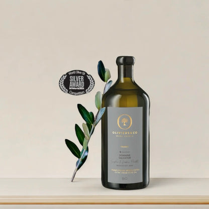 Flasche des Domaine Salvator Bio-Olivenöls aus Frankreich, Testsieger im Olivenöltest 2024, mit einem Silber-Award der NY World Olive Oil Competition. Die Flasche steht dekorativ neben einem Olivenzweig.