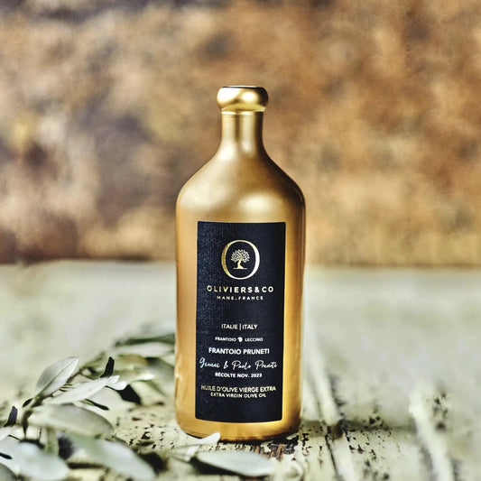 Elegante goldene Flasche Olivenöl aus Italien der Marke Oliviers & Co, platziert auf einem rustikalen Tisch mit Olivenzweigen im Vordergrund. Das schwarze Etikett trägt die Aufschrift "Frantoio Pruneti" und "Extra Virgin Olive Oil" sowie Angaben zur Herkunft aus Italien und das Erntedatum im November 2023. Die hochwertige Präsentation betont die Exklusivität des Olivenöls.