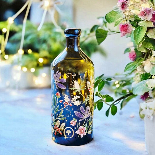 Limited Edition Olivenoelflasche aus Italien als Geschenk, kunstvoll mit floralen Motiven verziert, im Vordergrund eines weihnachtlichen Hintergrunds mit festlich dekorierten Zweigen.