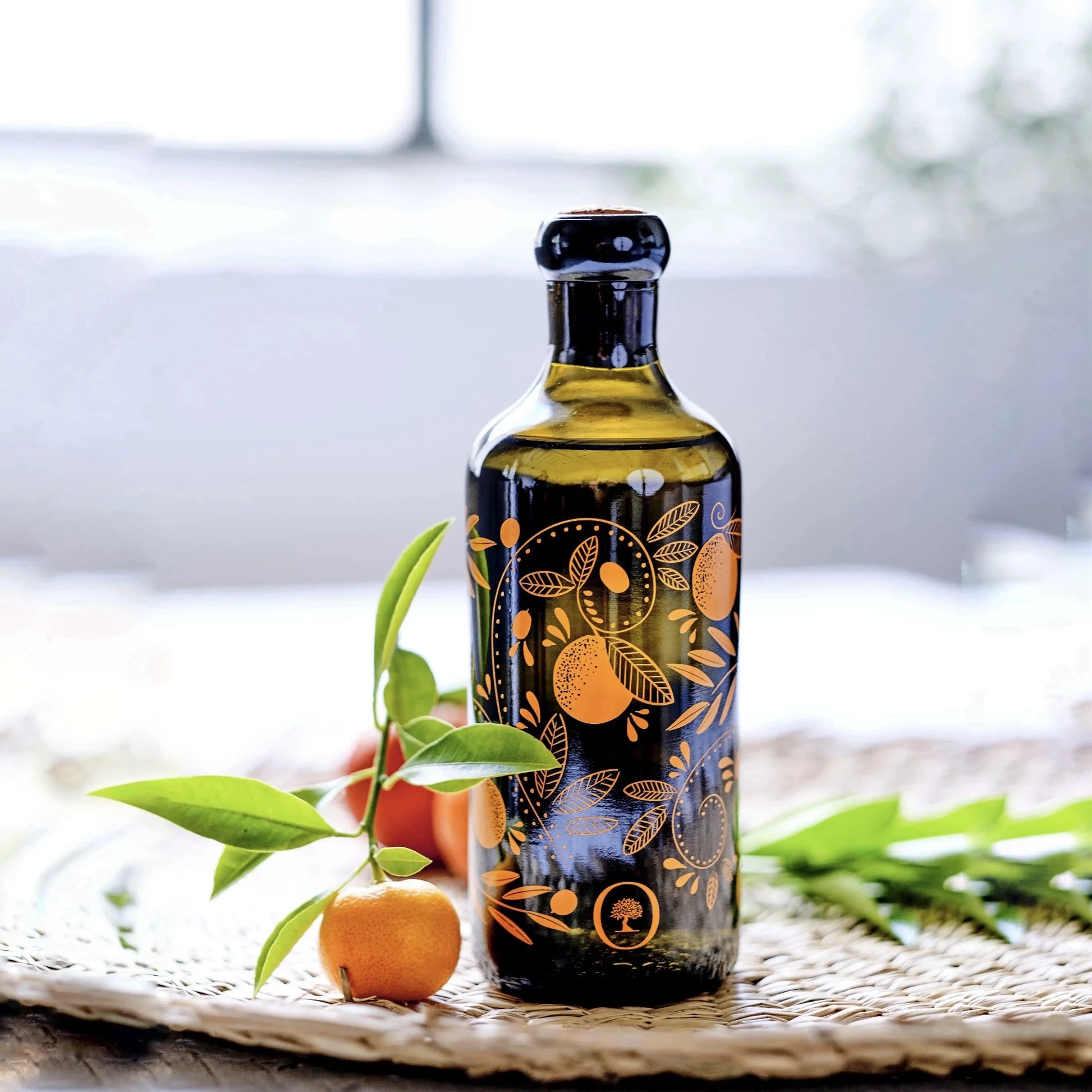 flasche mit olivenöl mit mandarinenaroma, verziert mit orangen mustern, auf einem geflochtenen untersetzer mit einer frischen mandarine und grünen blättern im vordergrund