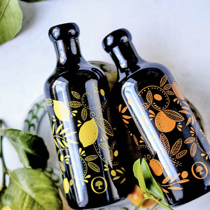 zwei flaschen mit olivenöl, eine mit zitronenaroma und die andere mit mandarinenaroma, verziert mit gelben und orangen mustern, auf einem teller mit grünen blättern im hintergrund