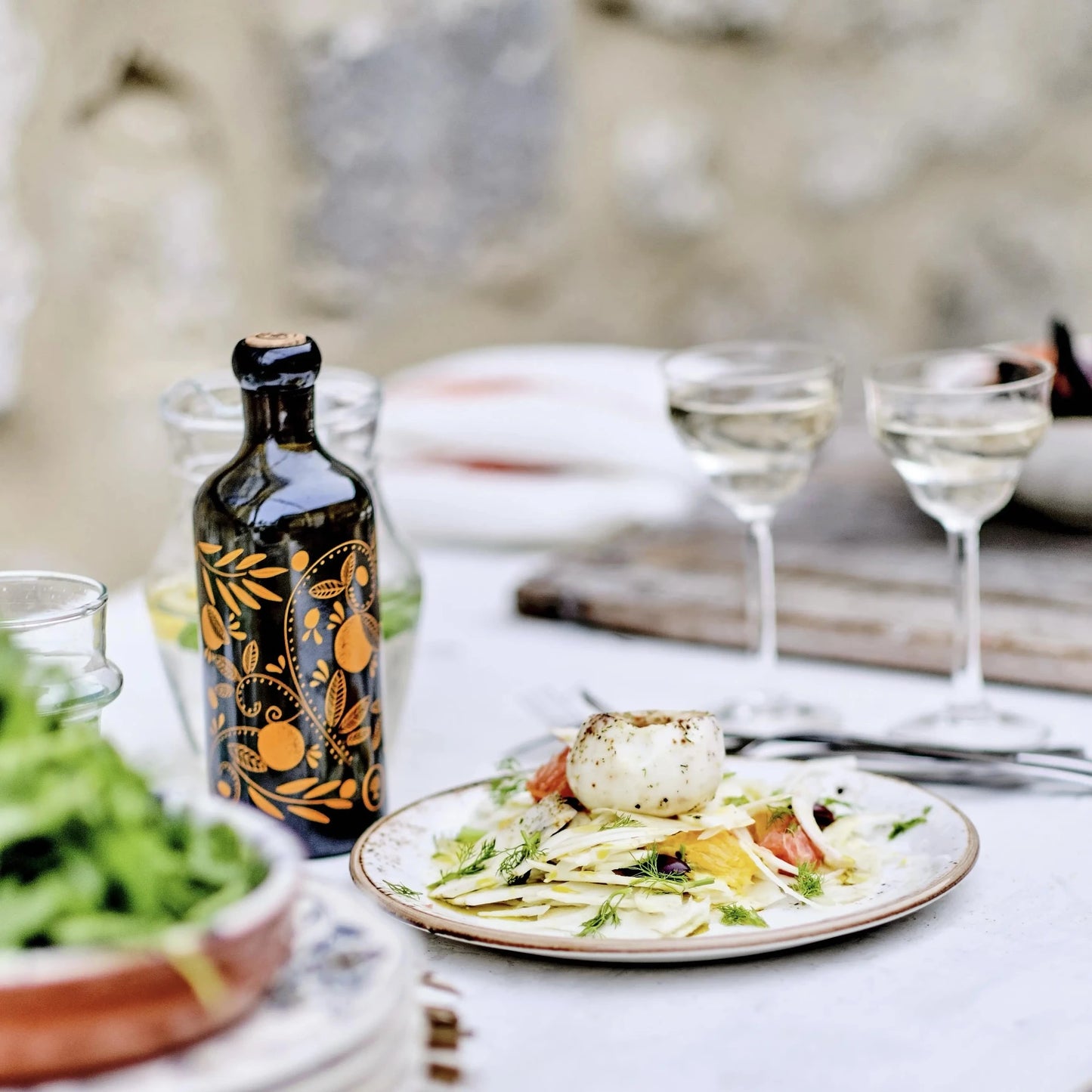 flasche mit olivenöl mit mandarinenaroma, verziert mit orangen mustern, auf einem gedeckten tisch neben einem teller mit einem gericht und zwei gläsern weißwein im hintergrund