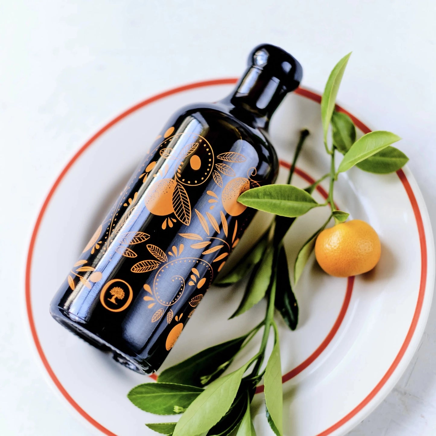 flasche mit olivenöl mit mandarinenaroma, verziert mit orangen mustern, liegend auf einem teller mit roten rand, neben einer frischen mandarine und grünen blättern