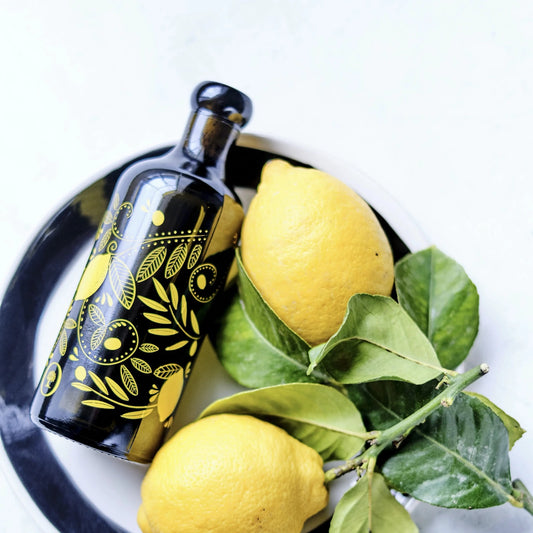 flasche olivenöl mit zitrone , verziert mit gelben mustern, liegend auf einem teller mit zwei frischen zitronen und grünen blättern