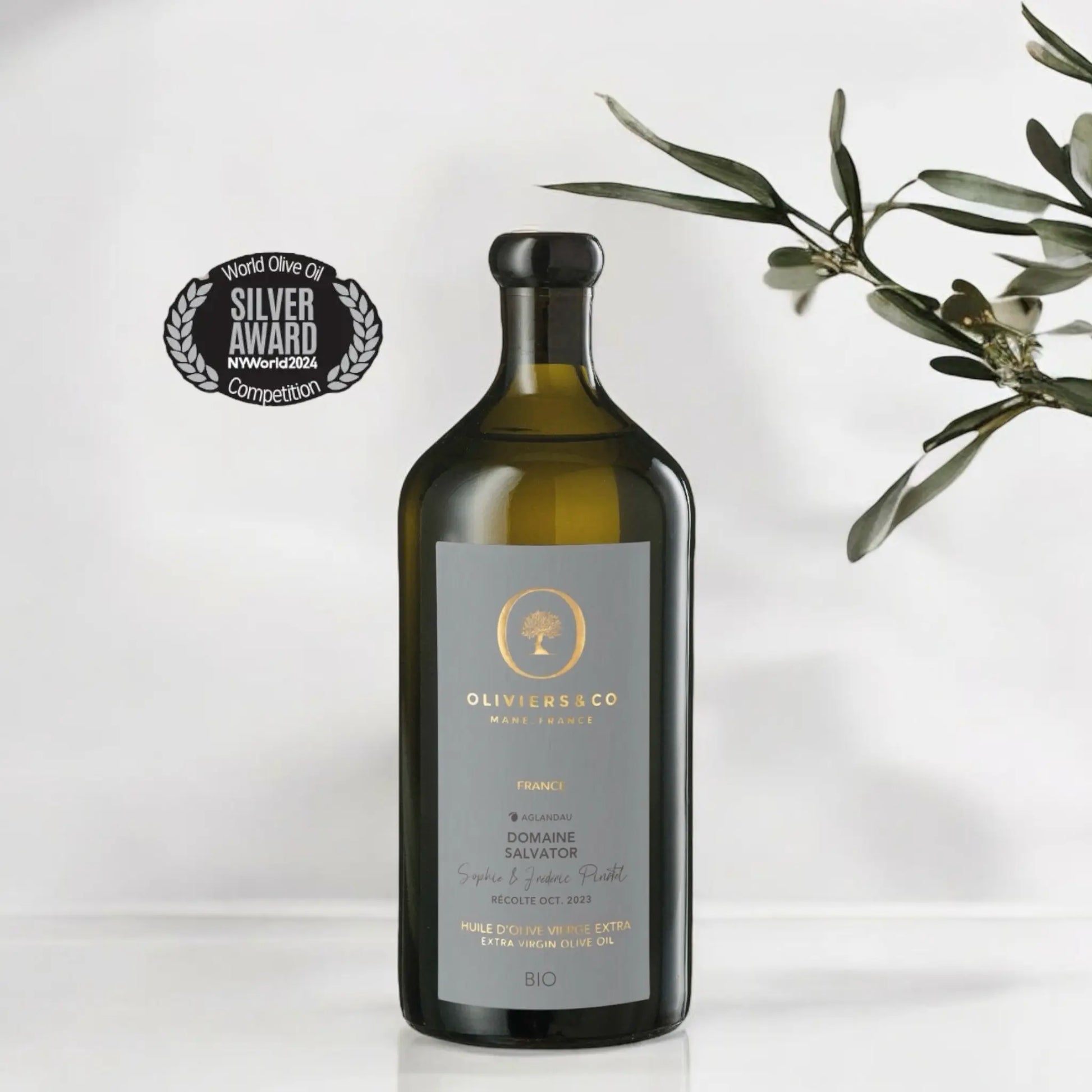 Flasche des Domaine Salvator Bio-Olivenöls aus Frankreich, Testsieger im Olivenöltest 2024, mit einem Silber-Award der NY World Olive Oil Competition. Die Flasche steht neben einem Olivenzweig auf einem hellen Hintergrund.