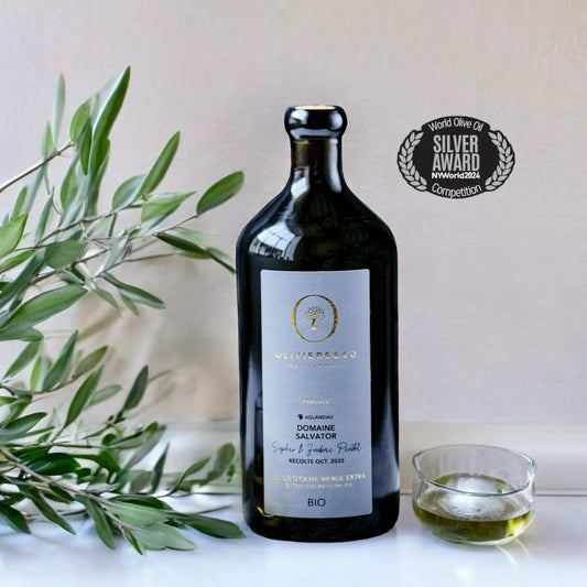 Flasche des Domaine Salvator Bio-Olivenöls aus Frankreich, Testsieger im Olivenöltest 2024, mit einem Silber-Award der NY World Olive Oil Competition. Die Flasche steht neben Olivenzweigen und einem Glas mit Olivenöl