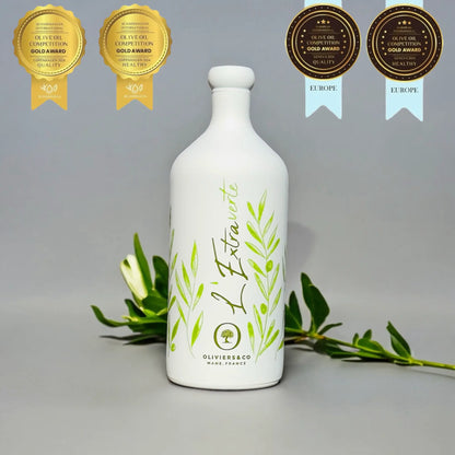 eine flasche testsieger olivenöl "l'extraverte" von oliviers & co, mane, france. umgeben von vier medaillen für qualität und gesundheit aus skandinavien und europa olivenöl test. im hintergrund olivenzweige
